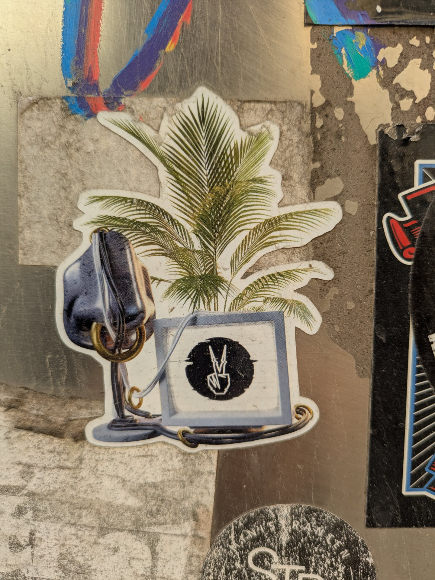 The sticker shows a vintage microphone next to a palm tree, below of which is a screen showing a symbol of a peace sign. This symbol appears to be in a circular frame. The elements are connected by metallic-looking rings and wires.