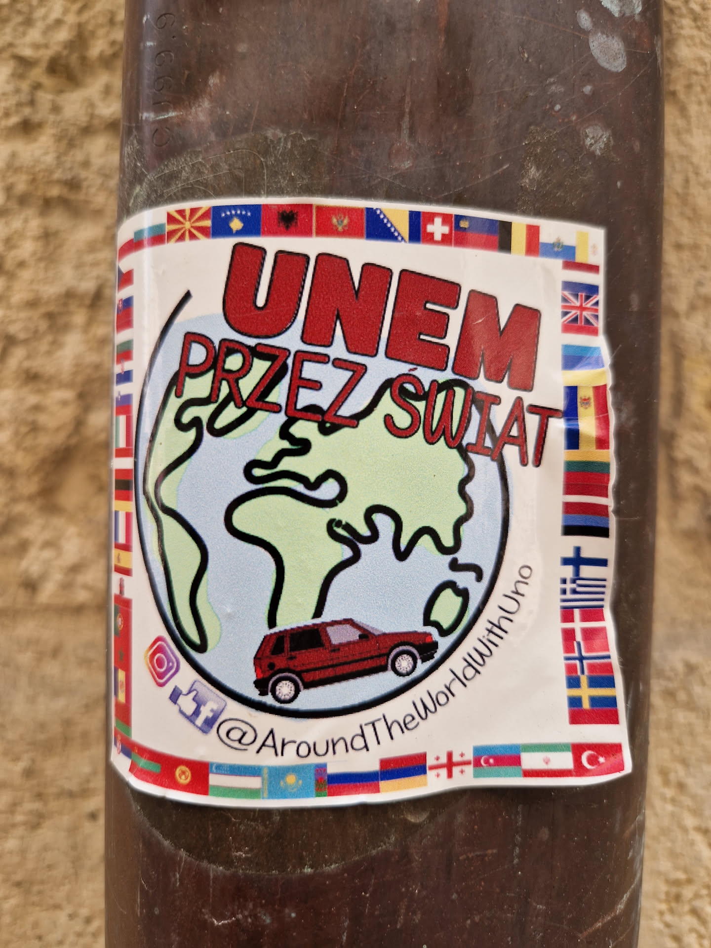 Sticker depicting a red car driving around the world, surrounded by various country flags. The text "UNEM PRZEZ ŚWIAT" is visible, along with a social media handle @AroundTheWorldwithUno.