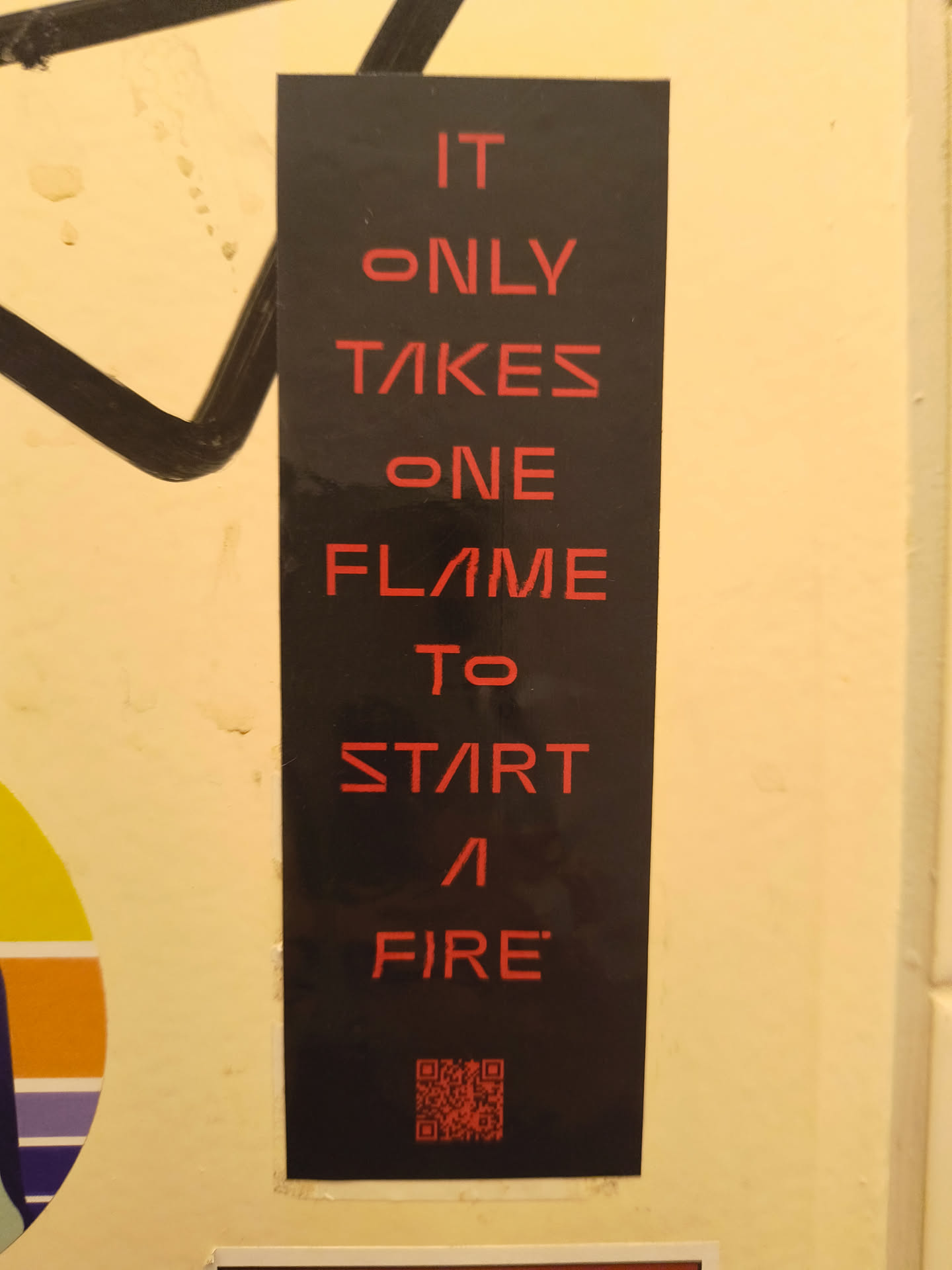 The sticker is a vertical rectangular shape with a matte black background. Bold, sans-serif text in a vibrant red color is centrally aligned, spelling out a motivational phrase. The font has a slightly futuristic or stylized appearance, particularly with the 'A's resembling an inverted 'V' and the 'E's having a broken middle bar. At the very bottom, a small, square QR code is present, also rendered in red against the black background. The sticker shows minor signs of wear, particularly at its edges, suggesting it's a physical object adhered to a wall.