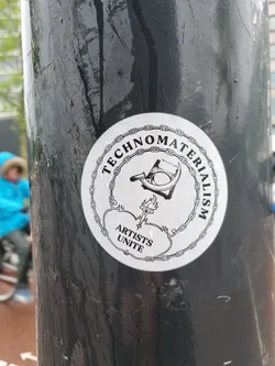A round sticker with a chain border. Inside, there's an illustration of a hammer and sickle with a torch, and the text "Technomaterialism" is written around the top. At the bottom, it says "Artists Unite".