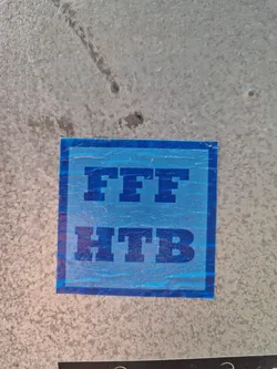 A light blue square sticker with the text "FFF HTB" in dark blue bold lettering. The sticker is affixed to a light grey, textured surface. The meaning of the acronym is unknown. 