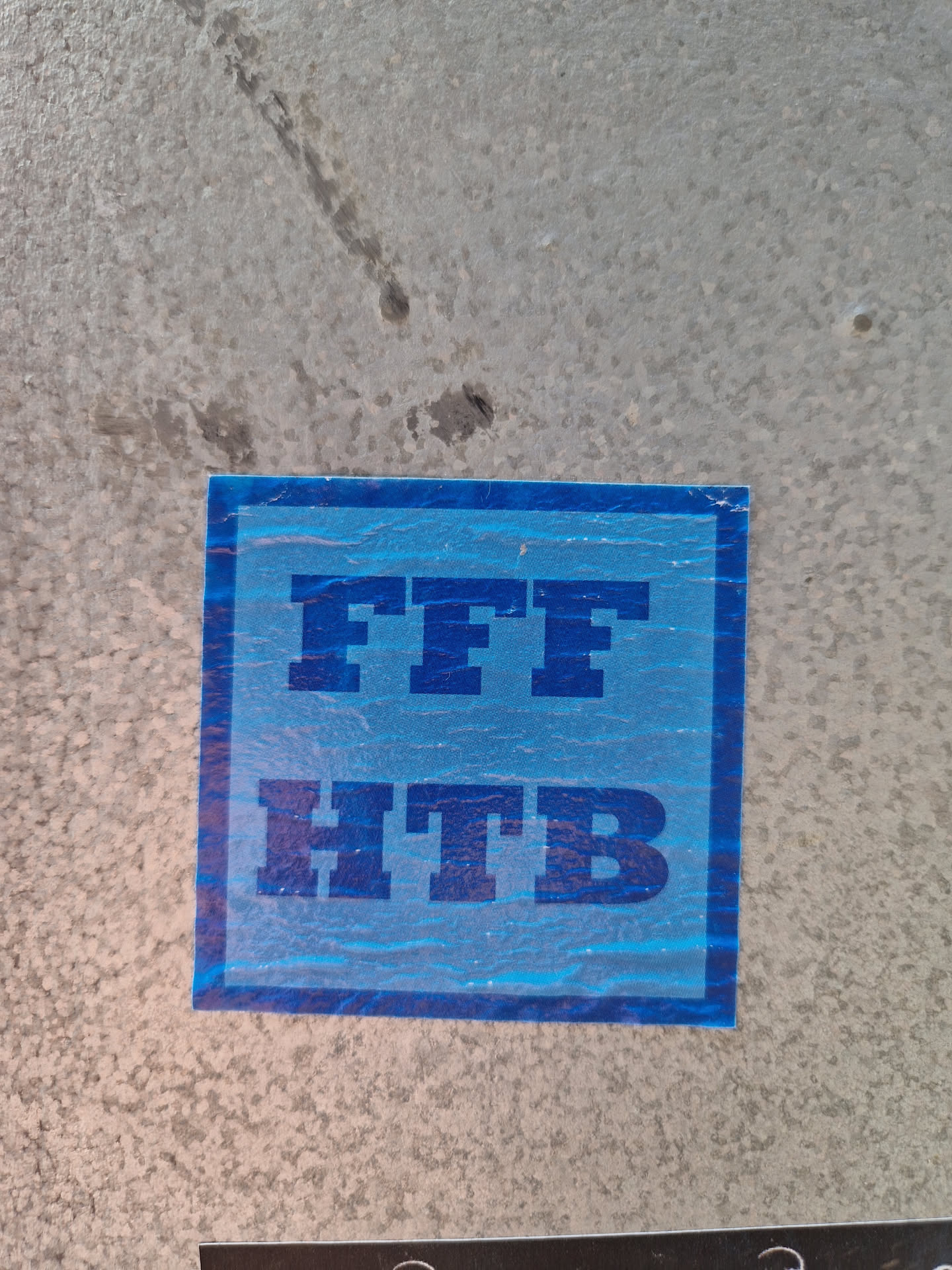 A light blue square sticker with the text "FFF HTB" in dark blue bold lettering. The sticker is affixed to a light grey, textured surface. The meaning of the acronym is unknown. 