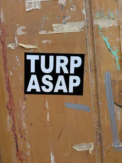 A rectangular sticker with a solid black background and prominent white, sans-serif capital letters. The text 'TURP' is on the top line, and 'ASAP' is on the bottom line, both centered. The sticker is affixed to a heavily weathered, rusty, and paint-splattered brown-orange surface, possibly a metal door or dumpster, creating a strong contrast between its clean design and the gritty environment.