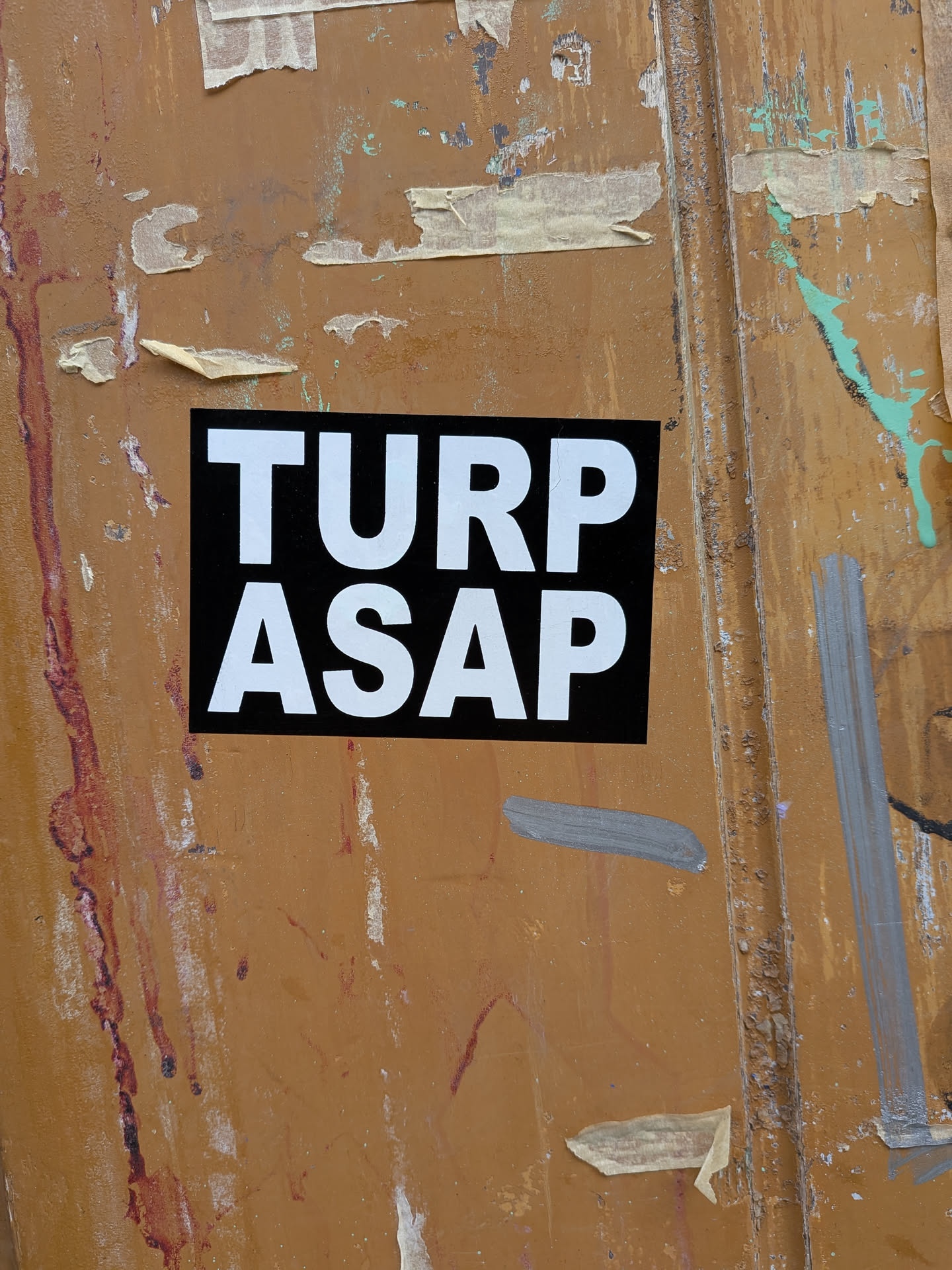 A rectangular sticker with a solid black background and prominent white, sans-serif capital letters. The text 'TURP' is on the top line, and 'ASAP' is on the bottom line, both centered. The sticker is affixed to a heavily weathered, rusty, and paint-splattered brown-orange surface, possibly a metal door or dumpster, creating a strong contrast between its clean design and the gritty environment.