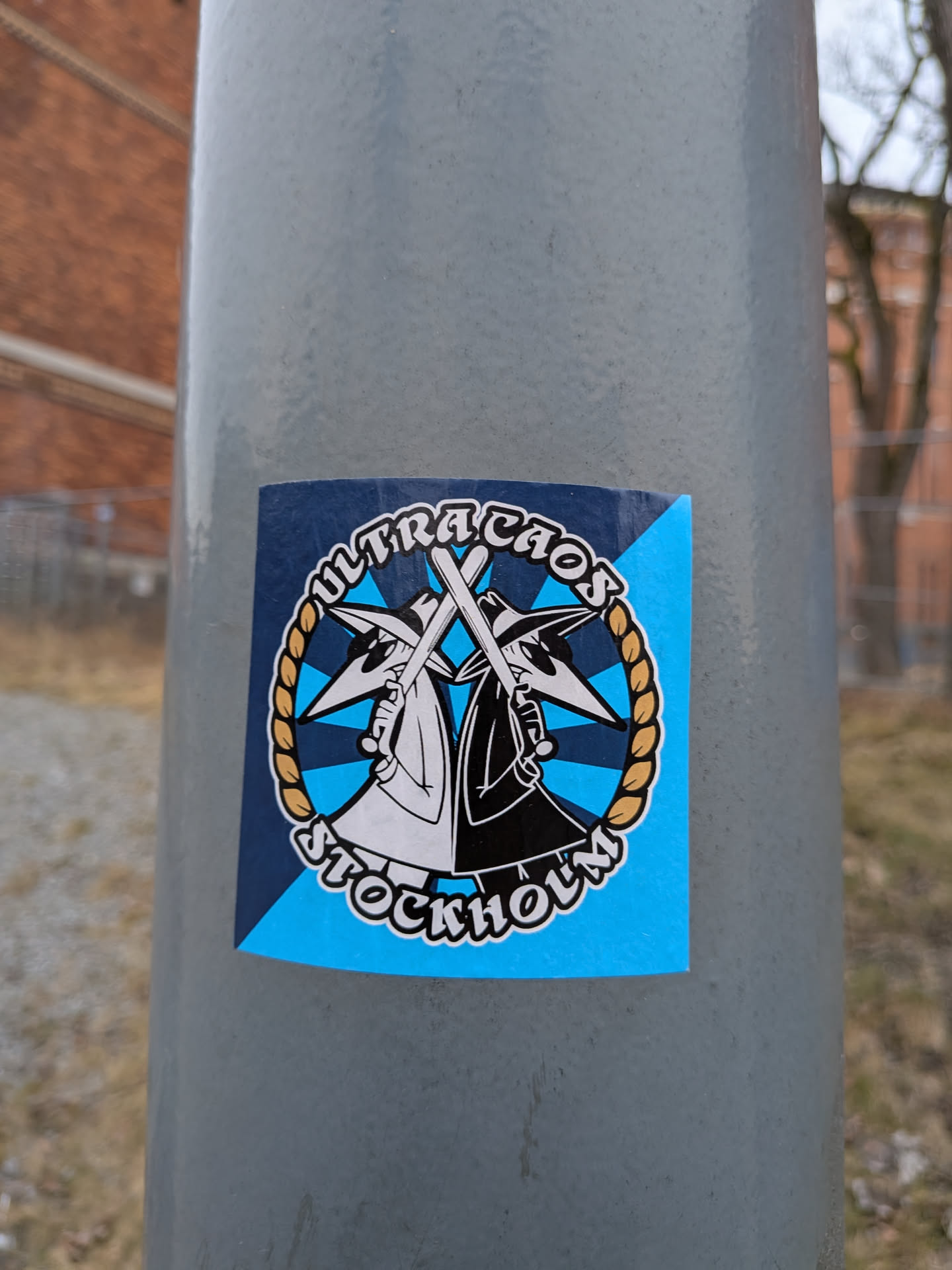 A circular sticker featuring two stylized figures, reminiscent of the 'Spy vs. Spy' characters, one in white and one in black, mirroring each other. Both figures wear pointed hats and dark sunglasses, and each holds a white baseball bat crossed in front. The background within the circle is light blue with dark blue star-like or triangular accents. A golden braided rope forms a border around the central figures. Arched text above reads 'ULTRACHAOS' and below reads 'STOCKHOLM', both in white with a black outline. The circular design is set against a larger square background that is diagonally split between dark blue and light blue. The sticker is applied to a grey pole.
