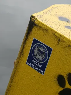A sticker on a yellow pole displays the Hertha BSC Berlin logo and the phrase "Gegen Rassismus" (Against Racism).