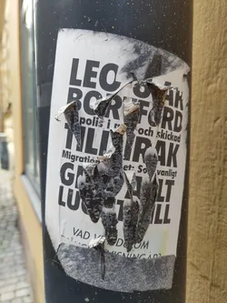 A heavily damaged sticker affixed to a dark grey pole.  Much of the text is illegible due to tearing and weathering.  Visible words include what appears to be 'Border', 'Police', 'Migration', and others in a foreign language. The sticker's original message is unclear due to its condition.