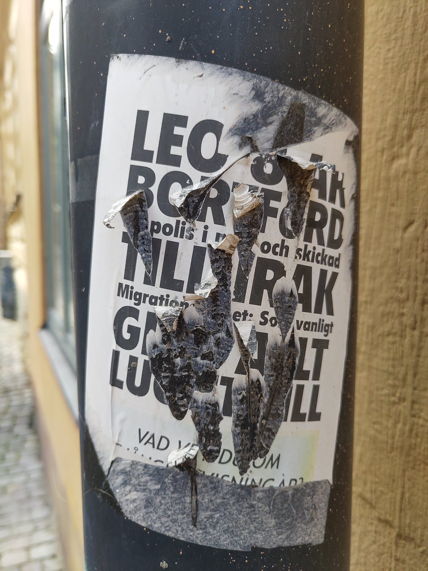 A heavily damaged sticker affixed to a dark grey pole.  Much of the text is illegible due to tearing and weathering.  Visible words include what appears to be 'Border', 'Police', 'Migration', and others in a foreign language. The sticker's original message is unclear due to its condition.