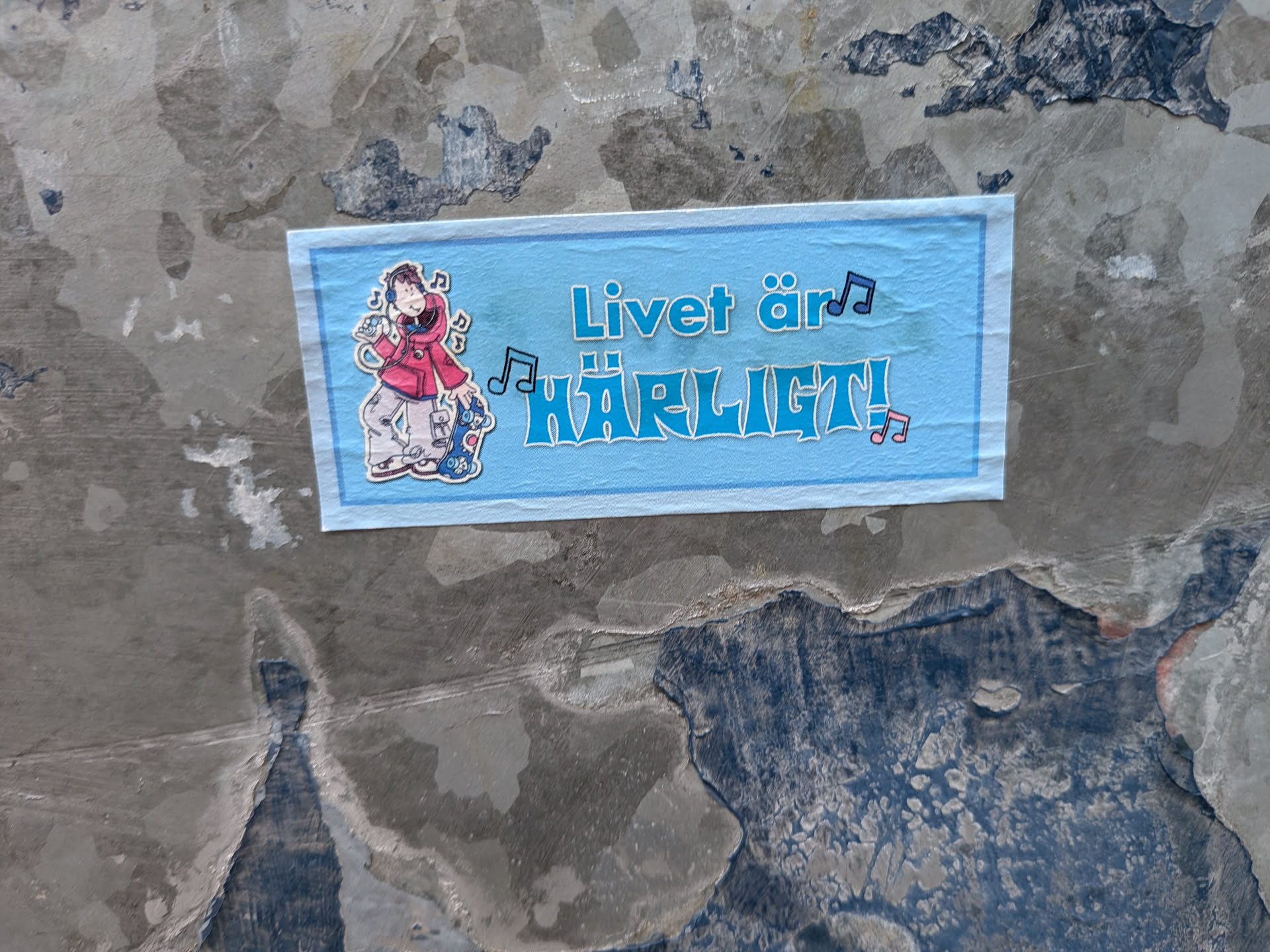 A rectangular light blue sticker with a darker blue border. On the left, it features a cartoon illustration of a person wearing headphones, a red hoodie, and grey pants, holding a skateboard. Musical notes float around the character. To the right, the text 'Livet är HÄRLIGT!' is written in blue letters with white outlines, interspersed with more musical notes. The sticker is pasted on a rough, weathered grey and blue wall.