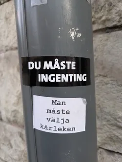 The image features two stickers on a gray pole. The upper sticker is black with white text that reads "DU MÅSTE INGENTING" (You have to nothing). Underneath it, there is a white sticker with the text:  "Man måste välja kärleken" (One must choose love) written in a typewriter-style font.