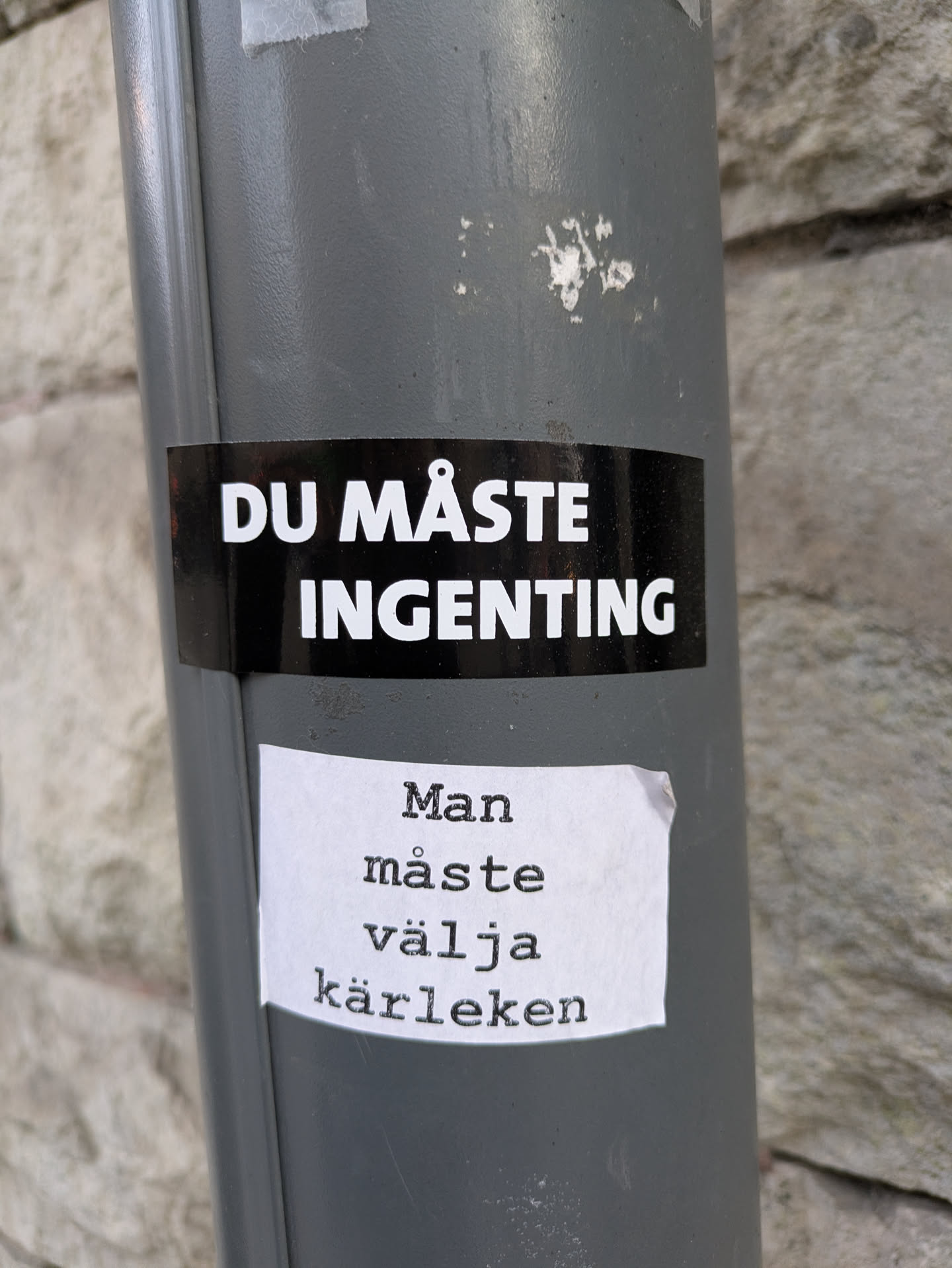 The image features two stickers on a gray pole. The upper sticker is black with white text that reads "DU MÅSTE INGENTING" (You have to nothing). Underneath it, there is a white sticker with the text:  "Man måste välja kärleken" (One must choose love) written in a typewriter-style font.