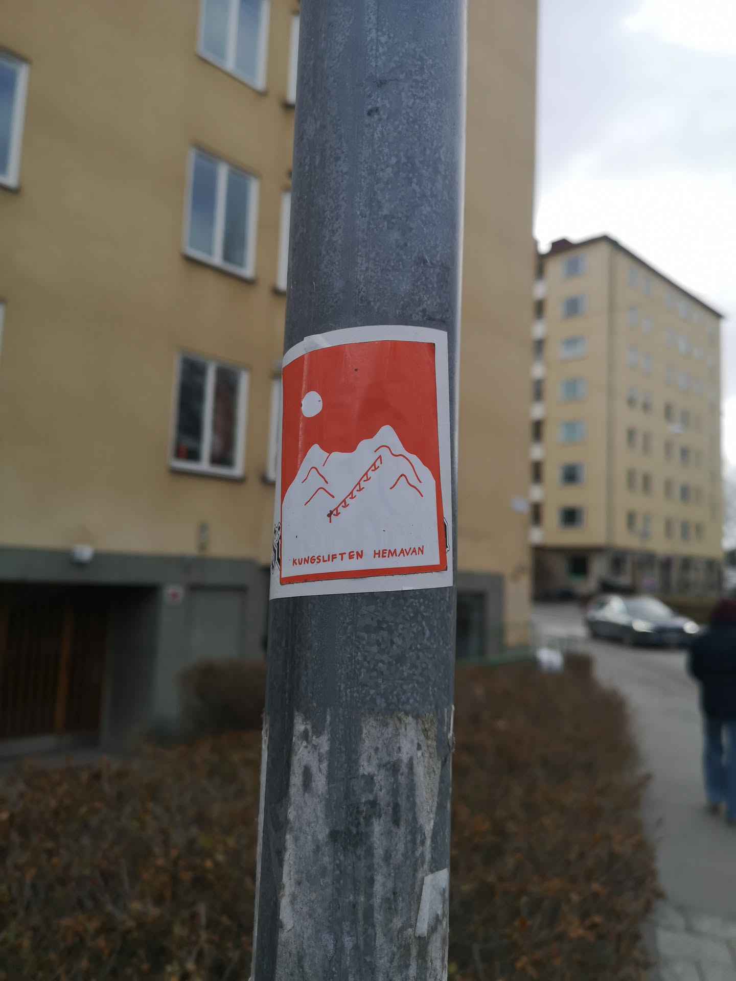 The sticker is rectangular with a clean white border, set against a weathered metal pole. The main design features a striking orange-red background. In the center, a white silhouette depicts a stylized mountain range with several peaks. Above the mountains, to the left, is a simple white circle, possibly representing the sun or moon. On the right side of the mountain, several red, jagged lines descend, suggesting ski tracks or a ski lift path. Below the mountain design, in the bottom left corner, the text "KUNGSLIFTEN HEMAVAN" is printed in red.