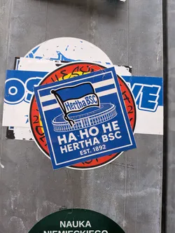 A square blue and white sticker representing the German football club Hertha BSC. It features the team's official waving flag emblem positioned above a stylized white line drawing of the Olympiastadion Berlin. Below the stadium illustration, bold white text reads 'HA HO HE HERTHA BSC EST. 1892'. The sticker is framed with a thin white interior border against a solid blue background.