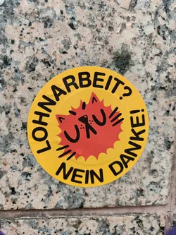 Street sticker Round yellow sticker with a red sun-like burst in the center. The burst has a stylized cat face and the text "Uru" inside.  The text around the circle says "LOHNARBEIT? NEIN DANKE!" which translates from German to: "Wage Labor? No Thanks!"