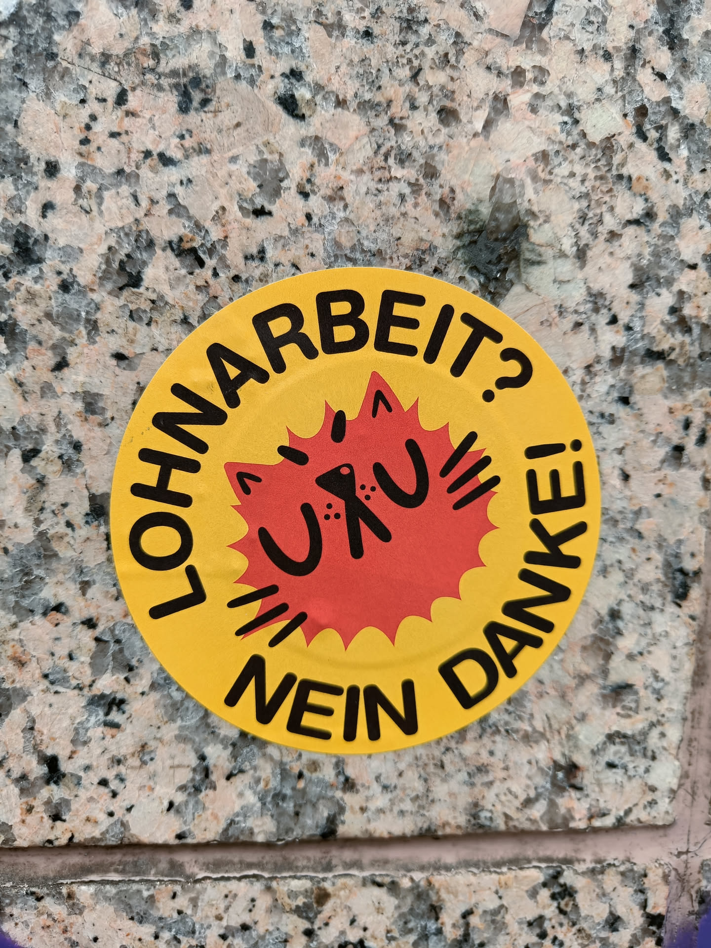 Round yellow sticker with a red sun-like burst in the center. The burst has a stylized cat face and the text "Uru" inside.  The text around the circle says "LOHNARBEIT? NEIN DANKE!" which translates from German to: "Wage Labor? No Thanks!"