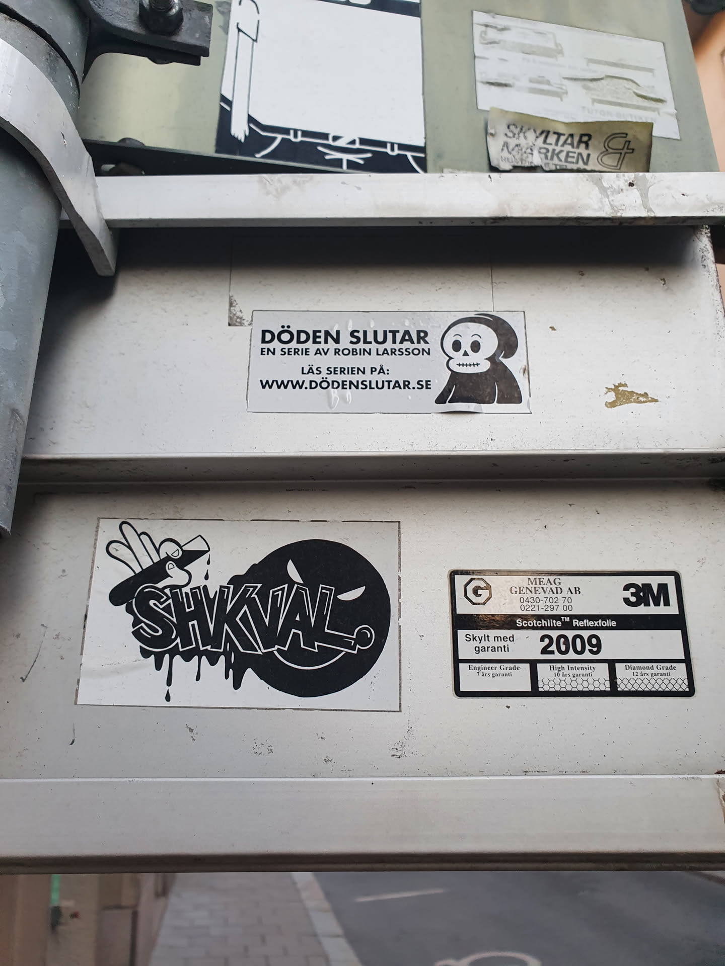 A black and white sticker with the word SHKVAL in graffiti style with a dripping effect. It is placed on a grey metallic surface along with other stickers and labels. 