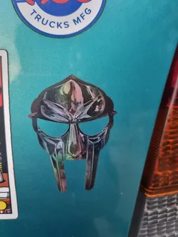 A sticker featuring the iconic mask of the rapper MF DOOM. The sticker has a holographic or iridescent effect, giving it a shiny, colorful appearance. The mask is depicted in its classic style, with its distinct features clearly visible.
