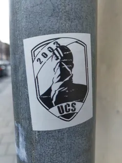Black and white sticker featuring a shield-shaped emblem. The emblem includes a stylized figure, the year 2003, and the letters UCS. The style is reminiscent of vintage or retro graphics.