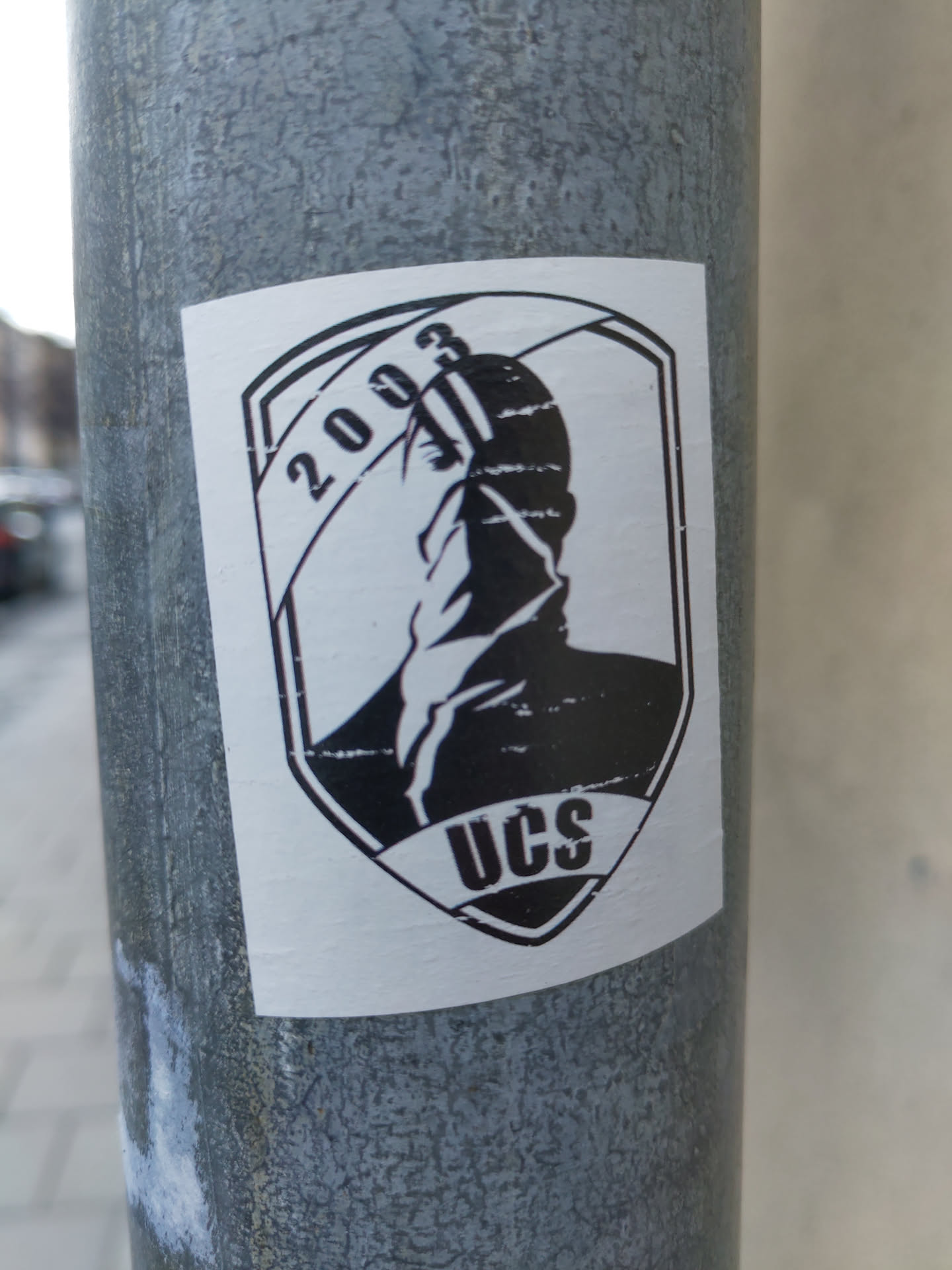 Black and white sticker featuring a shield-shaped emblem. The emblem includes a stylized figure, the year 2003, and the letters UCS. The style is reminiscent of vintage or retro graphics.
