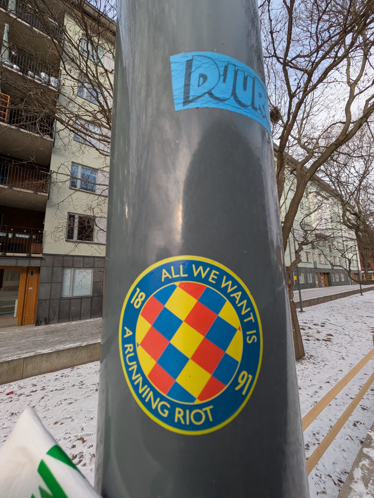 The sticker features a checkerboard pattern in red, yellow, and blue, with the text "ALL WE WANT IS 18 A RUNNING RIOT 91" encircling the design. The sticker is affixed to a gray post.