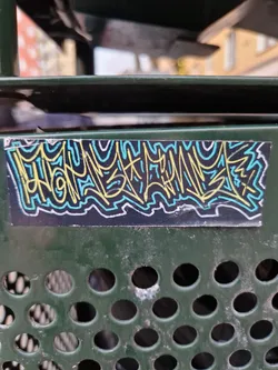 A sticker with an abstract graffiti design in yellow, light blue and teal on a black background is affixed to a dark-green perforated metal surface. The graffiti-style design is predominantly composed of stylized lines and curves.