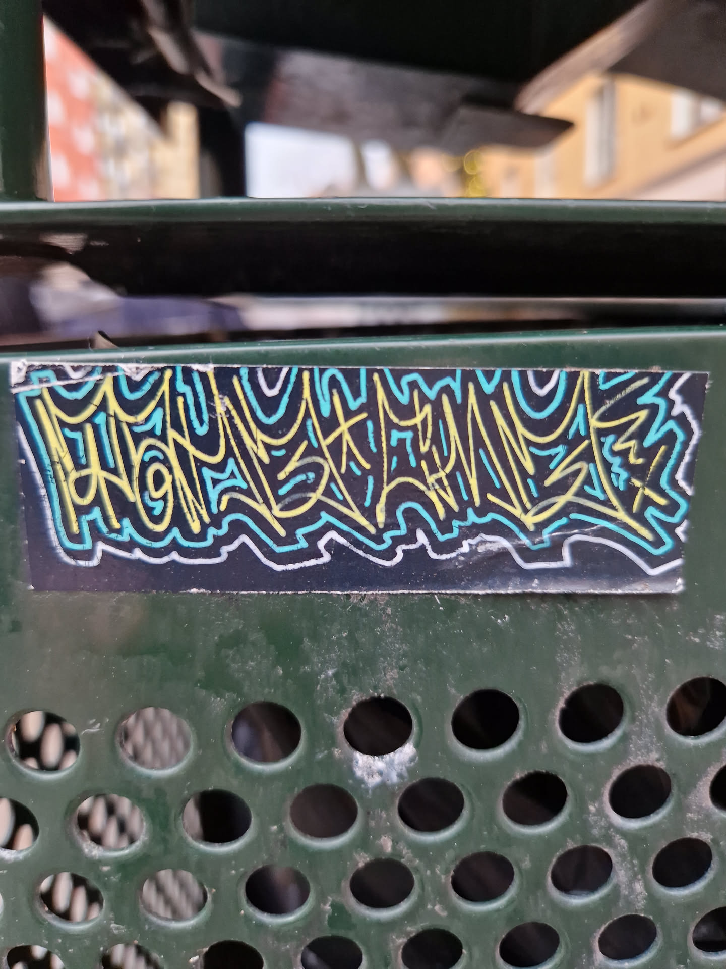 A sticker with an abstract graffiti design in yellow, light blue and teal on a black background is affixed to a dark-green perforated metal surface. The graffiti-style design is predominantly composed of stylized lines and curves.
