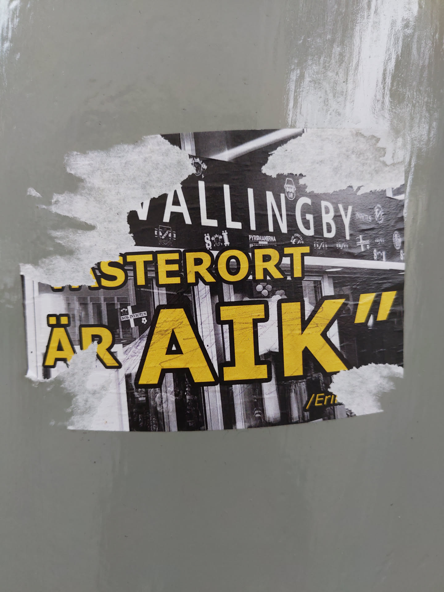 A sticker featuring a black and white image of a street scene with yellow text overlay that reads "VALLINGBY ÄR AIK". The text "VALLINGBY" is on top and smaller. The text "ÄR AIK" is larger and bolder, with the letters AIK being very prominent.  The sticker appears to be weathered or torn at the edges.