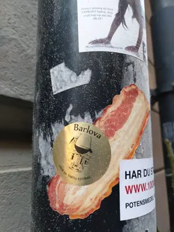 A gold-colored round sticker featuring the Barlova logo, which depicts a building and a wine glass, is displayed on a bacon-shaped sticker. The text on the Barlova sticker reads "Barlova" and "Pargi 28, Tartu, Estonia". Another sticker nearby says "HAR DU ST" and "WWW.1000POTENSMEDEL".