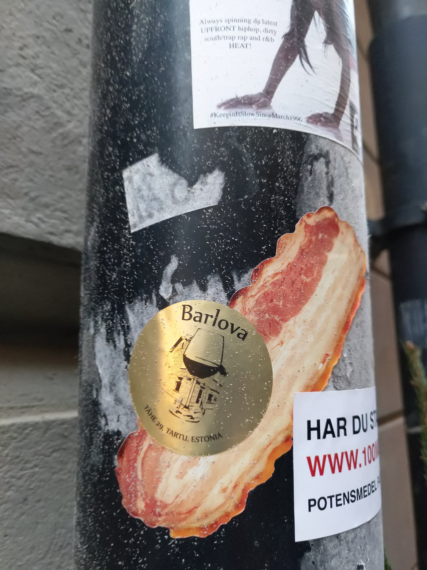 A gold-colored round sticker featuring the Barlova logo, which depicts a building and a wine glass, is displayed on a bacon-shaped sticker. The text on the Barlova sticker reads "Barlova" and "Pargi 28, Tartu, Estonia". Another sticker nearby says "HAR DU ST" and "WWW.1000POTENSMEDEL".