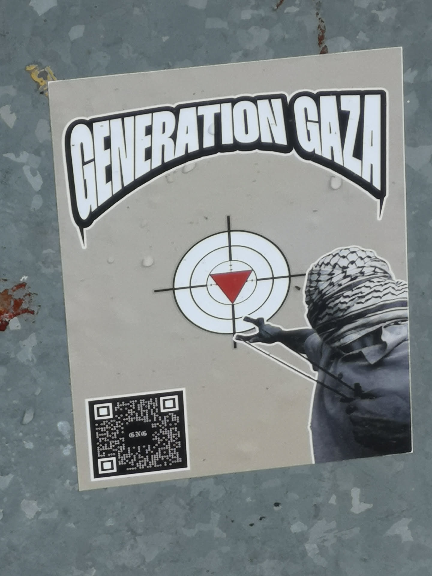 A rectangular sticker with a light grey background featuring a central graphic. At the top, the words "GENERATION GAZA" are displayed in large, bold, white letters with a thick black outline, forming an arch. Below this, a target symbol with concentric white and black circles and crosshairs is prominent, with a solid red inverted triangle in the bullseye. To the right, a grayscale figure, from the chest up, is depicted wearing a keffiyeh that covers their face, holding a slingshot and aiming towards the target. In the bottom left corner, there is a black and white QR code with the letters "GNG" visible in its center.