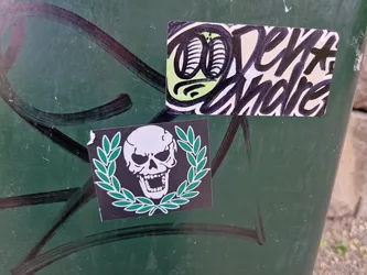 Close-up image of a sticker featuring a skull surrounded by a laurel wreath. The sticker is affixed to a dark green surface with some graffiti markings nearby. Another sticker with some stylized text is visible in the background. 