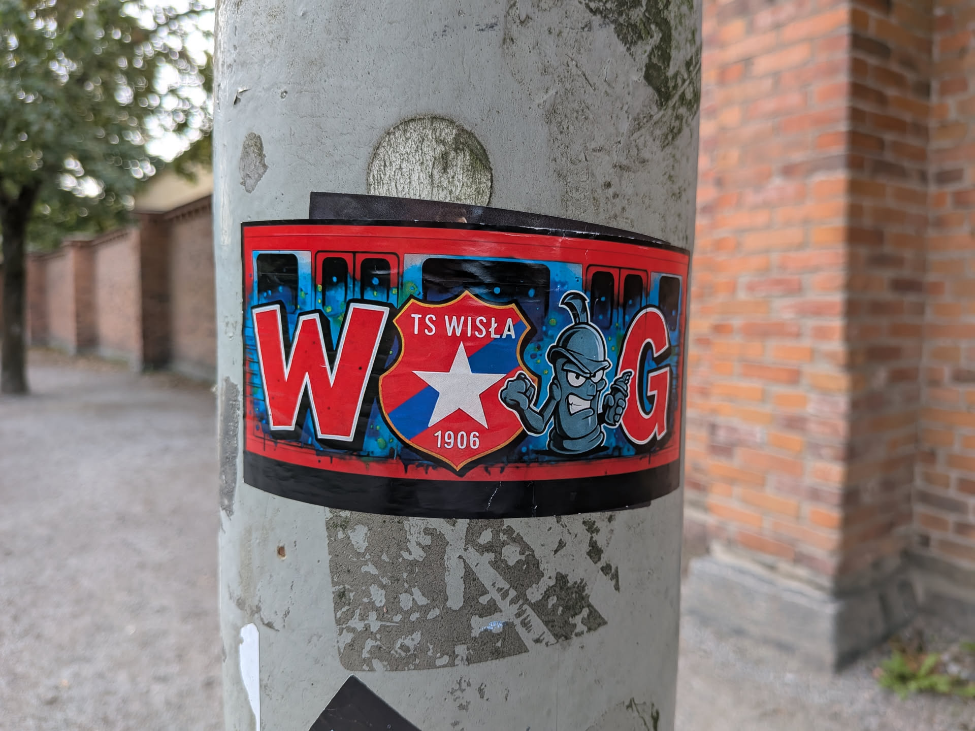 The sticker is rectangular with rounded corners and a red border. The background features a blue and black graffiti-style spray paint effect. On the left, a large red 'W' with a white outline is prominent. In the center, a shield-shaped emblem displays 'TS WISLA' at the top, a white star on a blue and red background, and '1906' at the bottom. To the right of the shield is a cartoonish, angry-looking character with a spray can, rendered in shades of blue and grey. A large red 'G' with a white outline is on the far right. The sticker appears to be slightly peeling at the top edge.