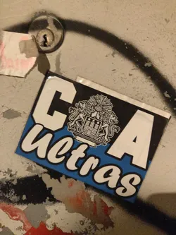 A sticker featuring the letters CA and the word "ultras" in a stylized font. A crest is prominently displayed above the text. The sticker is affixed to a weathered surface. 