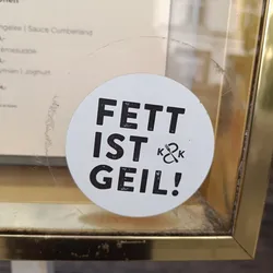 Round white sticker with black text. It says "FETT IST GEIL!" which translates from German to "Fat is cool!" There is a small logo with a fork and knife and the letters K&K under the text. The sticker is attached to a glass surface.