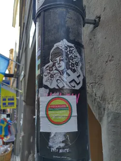 A black and white sticker featuring a stylized illustration of a woman with flowers in her hair, adorned with a cross. The sticker also includes the Instagram handle @danielflores7. Other stickers nearby include support information for Rojava, a region in northern Syria.
