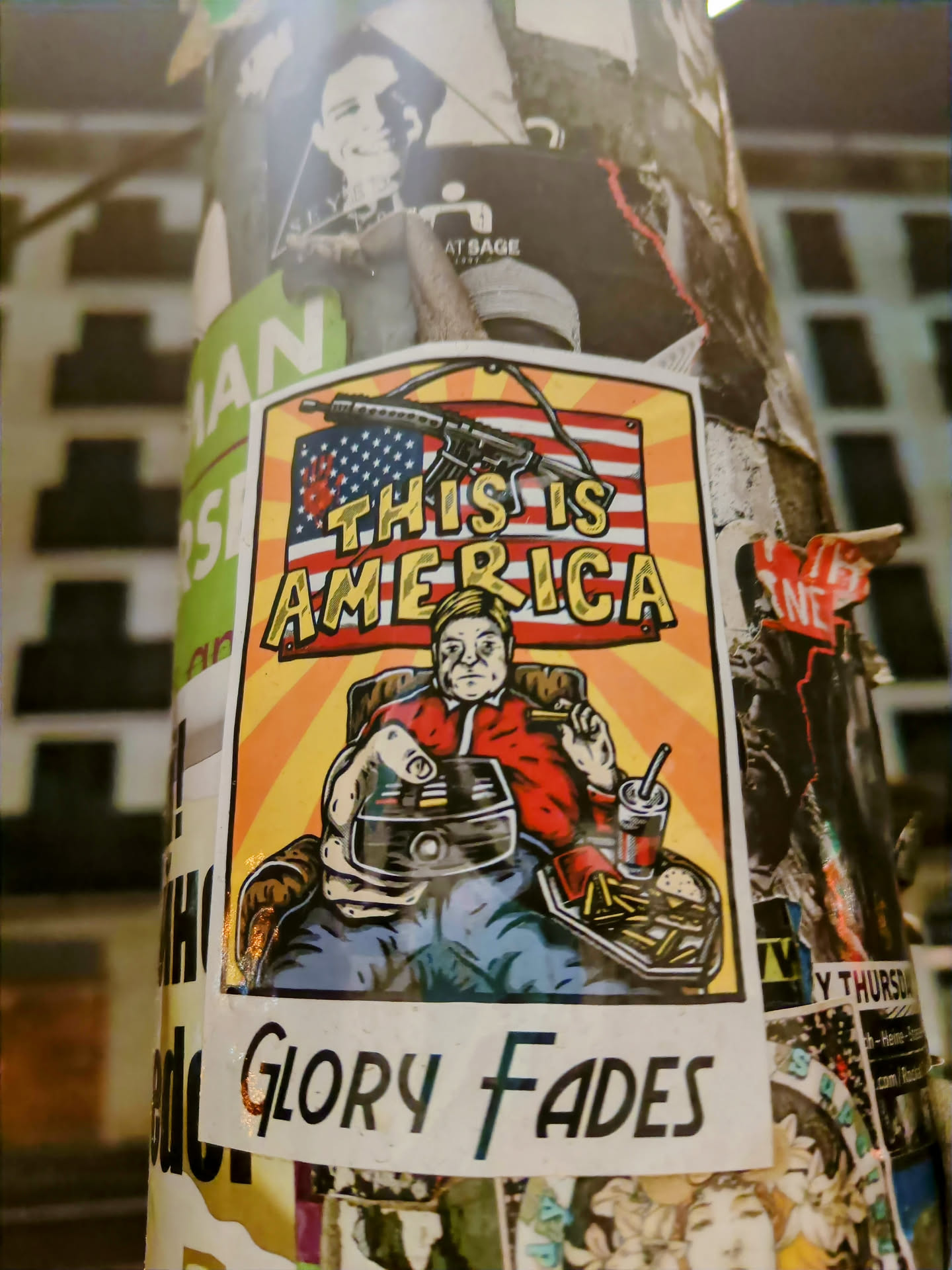 A sticker featuring a cartoon of a man sitting in a chair, holding a remote and enjoying fast food, with an American flag and a gun in the background. The text 'This is America' is prominently displayed, with the words 'Glory Fades' at the bottom.