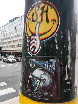 Two stickers on a pole. The top sticker is a smiley face with the letters SH and a thumbs up. The bottom sticker is a darker image of a foot in an Adidas shoe crushing a helmet. The background shows a street scene.