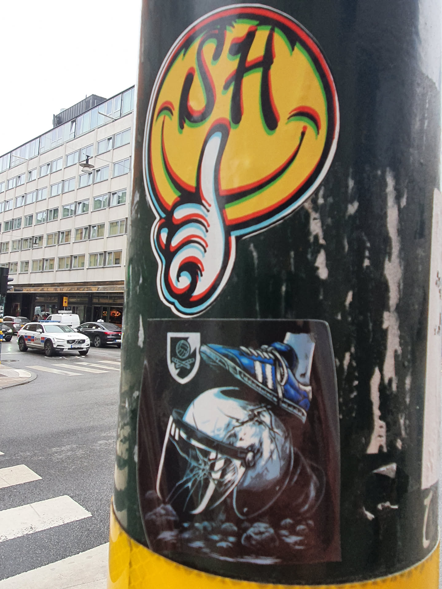 Two stickers on a pole. The top sticker is a smiley face with the letters SH and a thumbs up. The bottom sticker is a darker image of a foot in an Adidas shoe crushing a helmet. The background shows a street scene.