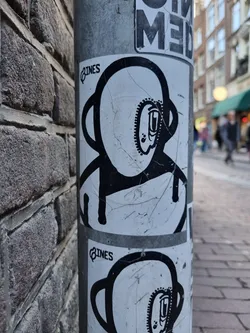 A worn-out sticker featuring a simple, black and white illustration of an astronaut-like character. The character has a round head with a smaller circular detail inside, and a simple body. It's been stuck to a metal pole, resulting in some creases and scratches on the sticker surface.  The word 'LINES' is visible in a small font near the top of the sticker.