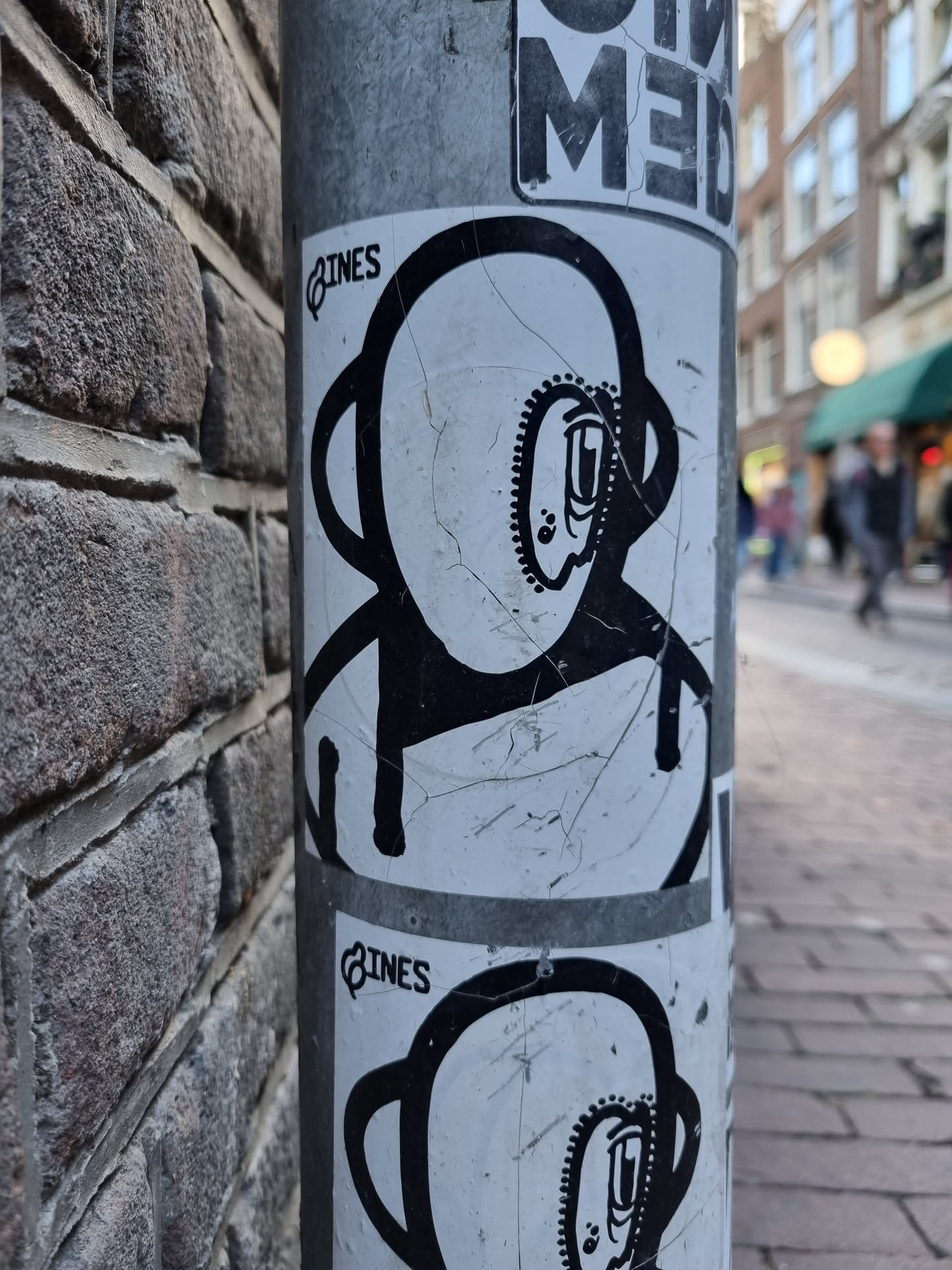 A worn-out sticker featuring a simple, black and white illustration of an astronaut-like character. The character has a round head with a smaller circular detail inside, and a simple body. It's been stuck to a metal pole, resulting in some creases and scratches on the sticker surface.  The word 'LINES' is visible in a small font near the top of the sticker.