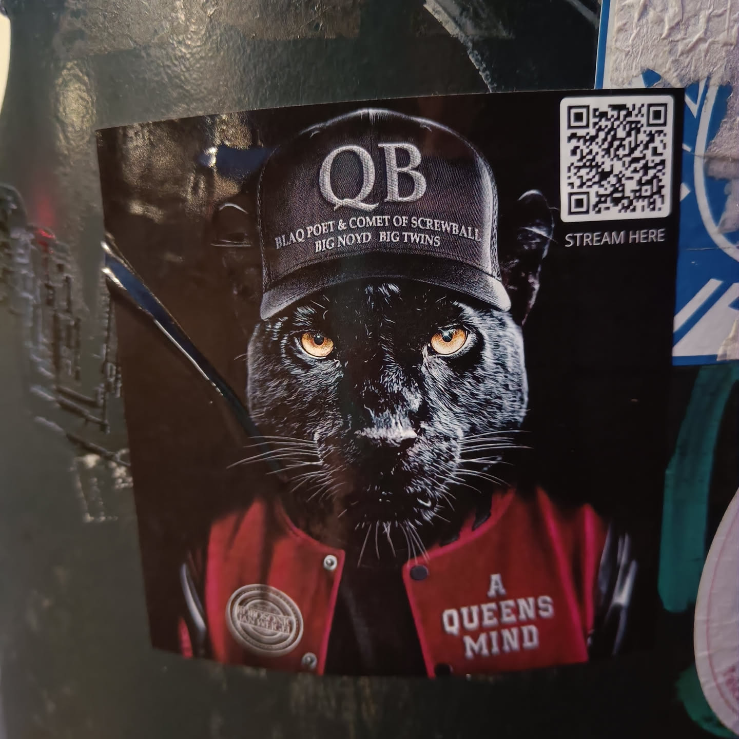 A sticker featuring a black panther wearing a baseball cap with the letters "QB" and the names "Blaq Poet & Comet of Screwball, Big Noyd, Big Twins" on it. It's also wearing a red varsity jacket with the words "A Queens Mind" on the back. There is a QR code in the corner.