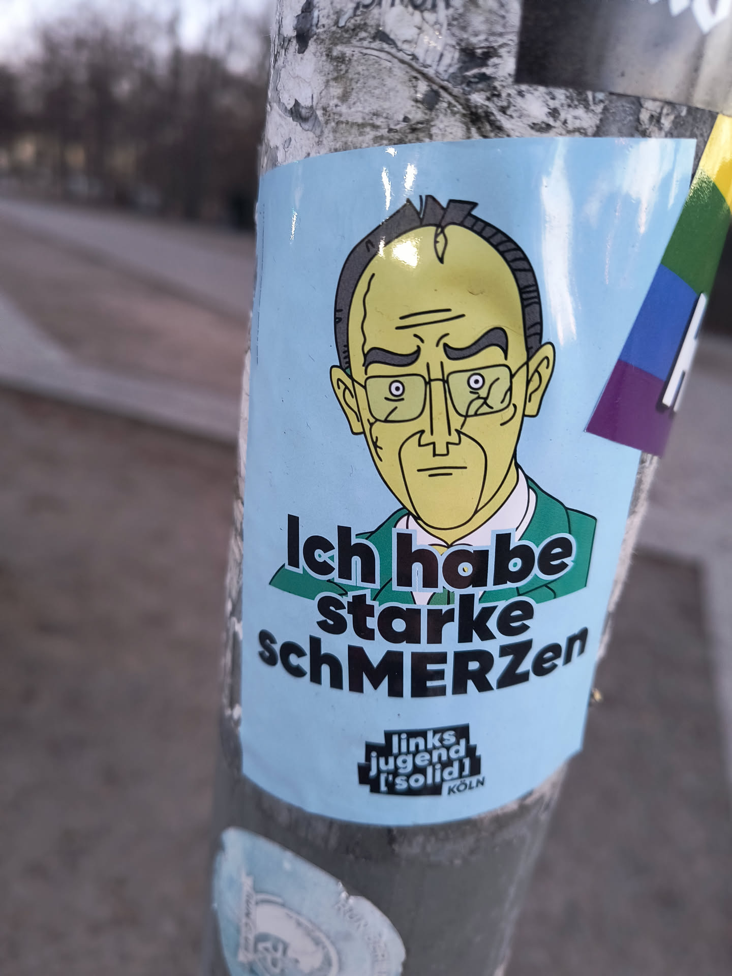 The sticker features a cartoon caricature of a man, likely Friedrich Merz, with a pale yellow-green skin tone, dark hair, and rectangular glasses. He wears a dark green suit jacket over a white shirt, depicted from the chest up. His expression is one of weariness or pain, with heavy-lidded eyes and a downturned mouth. The background of the sticker is a solid light blue. Below the figure, in bold black letters, is the German phrase "Ich habe starke schMERZen" (I have strong pain), with 'MERZ' capitalized. At the bottom, there is a black rectangular logo with white text that reads "links jugend ['solid] KÖLN". The sticker is applied to a metal pole and shows some minor reflections and slight wear.