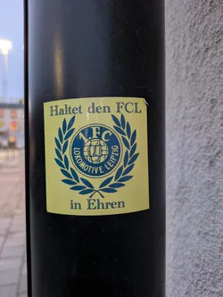 A sticker showing support for F.C. Lokomotive Leipzig.  The sticker is yellow with a blue and green emblem featuring the team's logo and a laurel wreath. The text "Haltet den FCL in Ehren" (Keep the FCL in Honor) is displayed above and below the emblem.