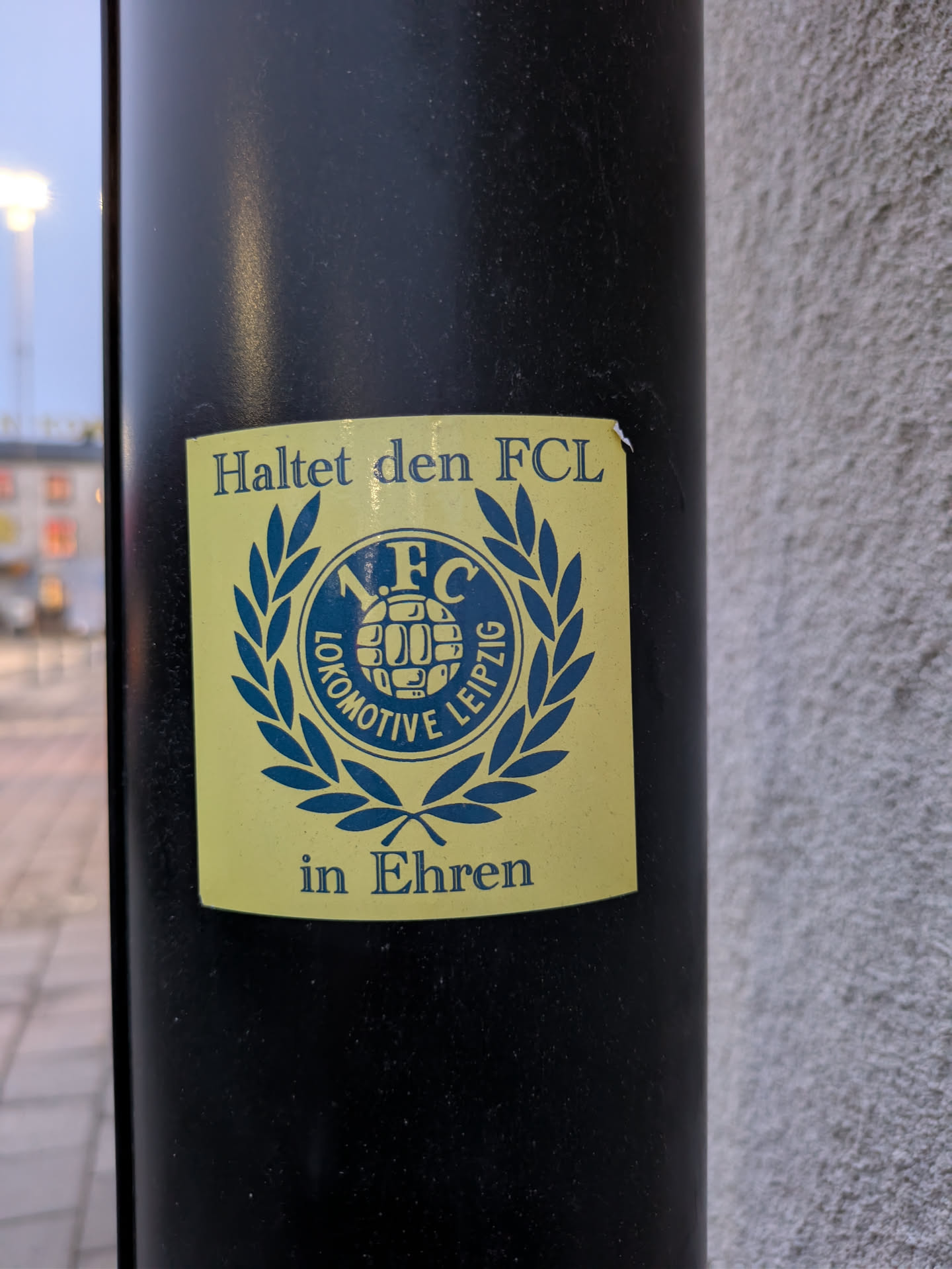 A sticker showing support for F.C. Lokomotive Leipzig.  The sticker is yellow with a blue and green emblem featuring the team's logo and a laurel wreath. The text "Haltet den FCL in Ehren" (Keep the FCL in Honor) is displayed above and below the emblem.