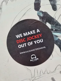 Round black and red sticker with the text "We make a disc jockey out of you" and a website address. There is a logo at the bottom that says DJ Les Amsterdam.