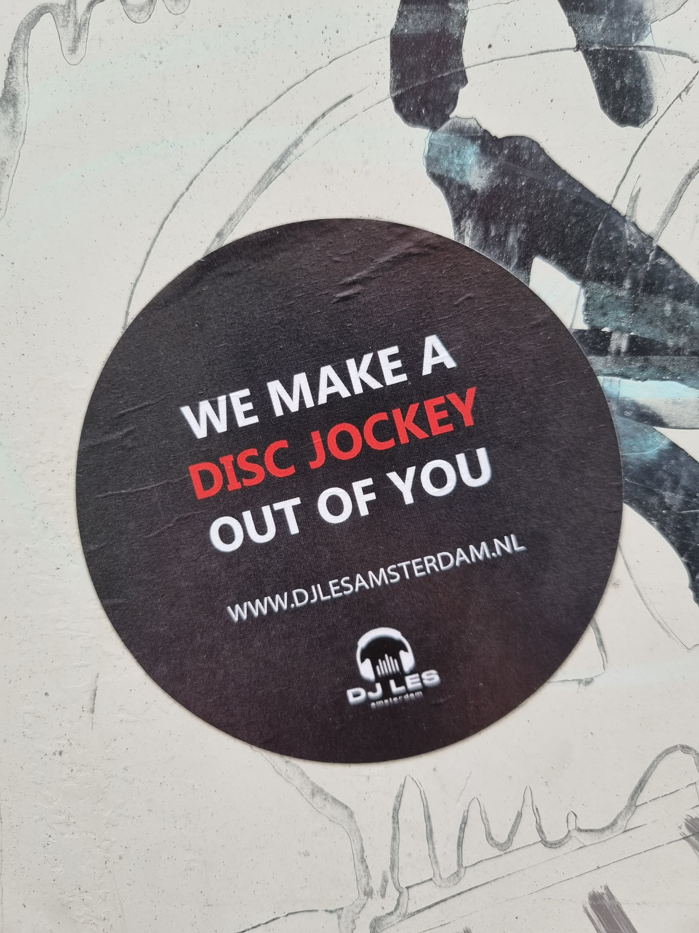 Round black and red sticker with the text "We make a disc jockey out of you" and a website address. There is a logo at the bottom that says DJ Les Amsterdam.