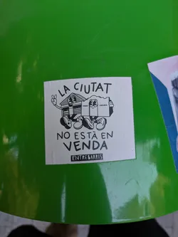 This sticker features a cartoon drawing of a house character with a peace sign giving hand and a box character that holds a sign and wearing shoes. The text on the sticker reads "LA CIUTAT NO ESTA EN VENDA" and "ENTREBARRIS". It seems to be against selling the city. The image is on a green surface.