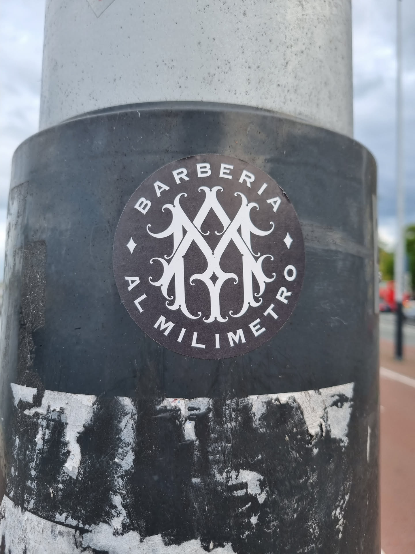A round black and white sticker featuring the logo of Barberia Al Milimetro. The logo is a monogram with stylized letters and ornate details. The text 'BARBERIA' and 'AL MILIMETRO' are written around the monogram. The sticker is affixed to a dark-grey pole.