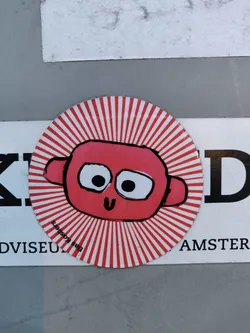 Round sticker featuring a cartoonish pink monster with large eyes and a small smile. The monster is set against a white background with radiating red stripes, creating a sunburst effect. The sticker appears to be hand-drawn or digitally created to mimic a hand-drawn style. There's also some small text visible on the sticker, but it's not clear enough to read.