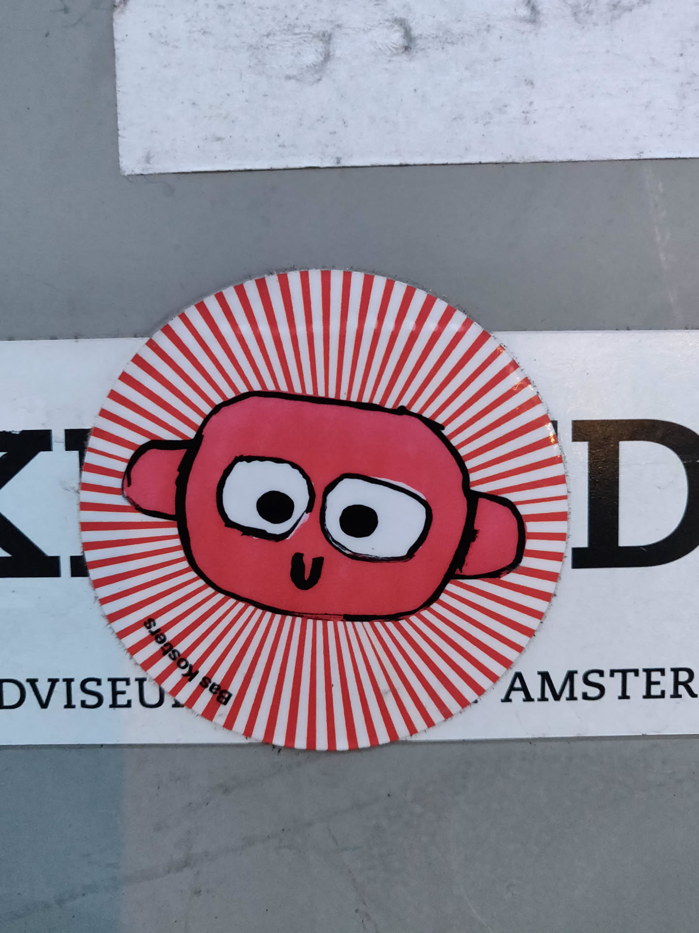 Round sticker featuring a cartoonish pink monster with large eyes and a small smile. The monster is set against a white background with radiating red stripes, creating a sunburst effect. The sticker appears to be hand-drawn or digitally created to mimic a hand-drawn style. There's also some small text visible on the sticker, but it's not clear enough to read.